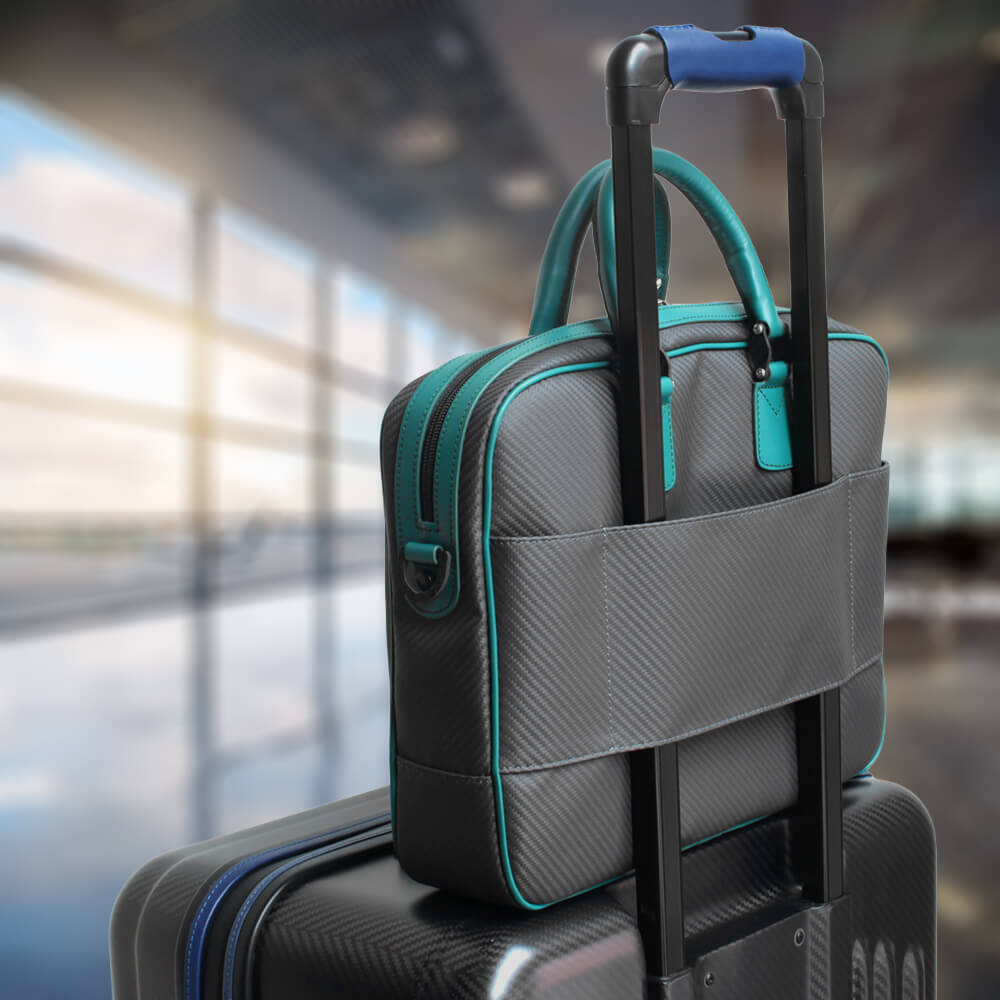 A small business bag in soft carbon material, featuring teal accents, positioned on top of a suitcase against a blurred airport background.